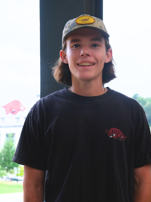 An image of Alex, a young male with chin length hair, smiling wide in front of Razorback Stadium