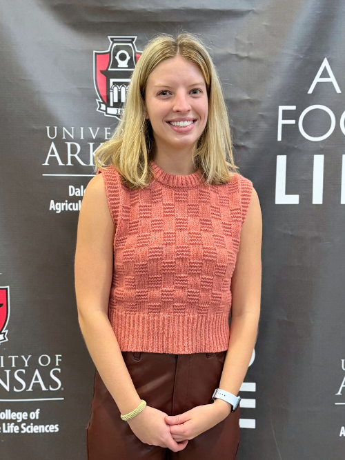 An image of Anna, a young woman with shoulder length blond hair. Anna is smiling wide with her hands clasped in front of her while standing in front of a black and white Bumpers College backdrop.