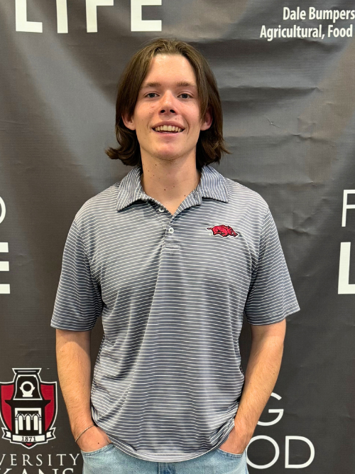 An image of Alex, a young male with chin length hair, smiling wide in front of a black and white Bumpers College backdrop.