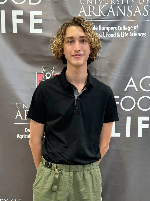 An image of Braeden, a young person with longer curls is smiling at the camera with their hands clasped behind their back. Braeden is standing in front of a black and white Bumpers College backdrop. 
