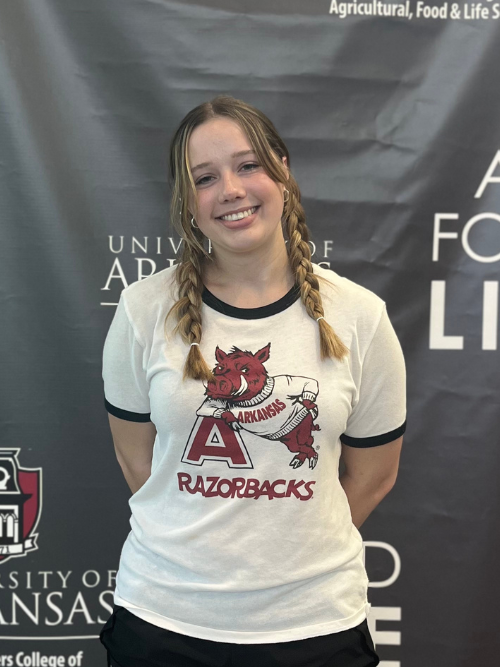 Image of Delaney with blonde braided pigtails smiling at the camera with her arms clasped behind her, wearing a vintage University of Arkansas white, red, and black tee.