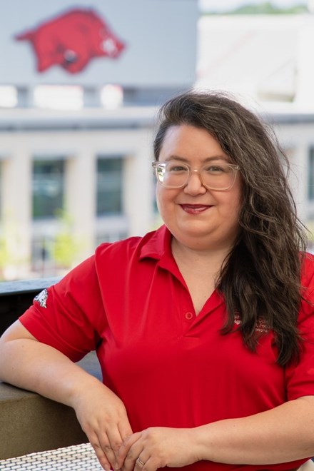 Headshot of a white, hispanic woman with long brown curly hair smiling at the camera. She is wearing a red Razorback polo, clear glasses, red lipstick, and standing in front of the Razorback stadium.