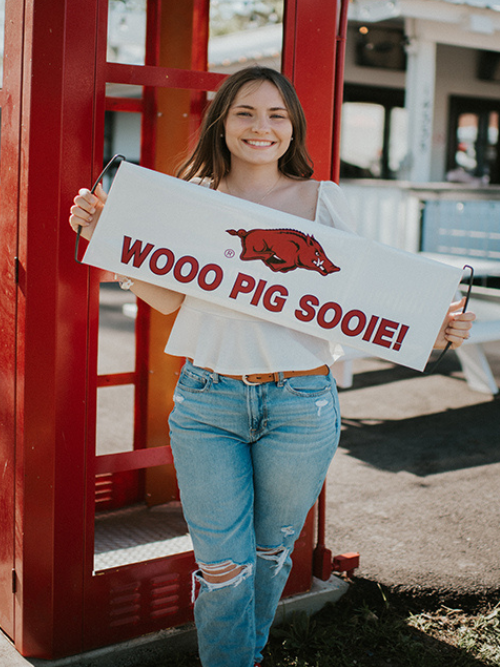 Image of Kerri smiling in front of a red telephone booth holding a sign that says woo pig sooie.