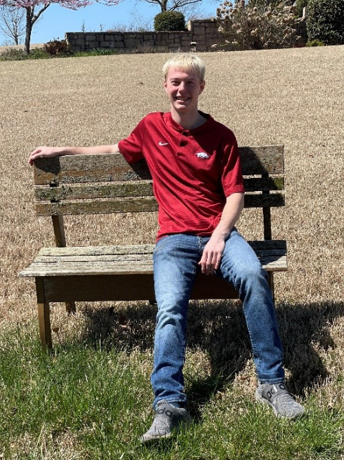 Image of Kole sitting outdoors on a garden bench, smiling and wearing a red shirt