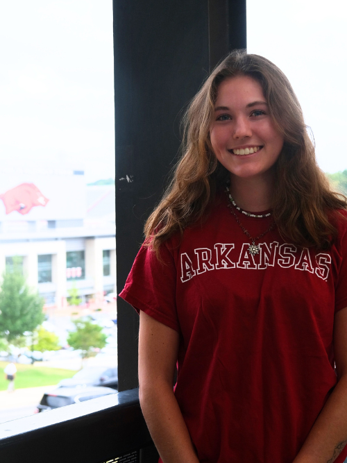 An image of Natalie, a young woman with shoulder length wavy light brown hair, smiling wide in front of Razorback Stadium