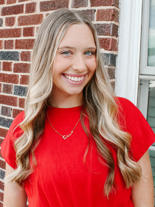 Image of Olivia standing in front of a brick wall smiling in a red blouse