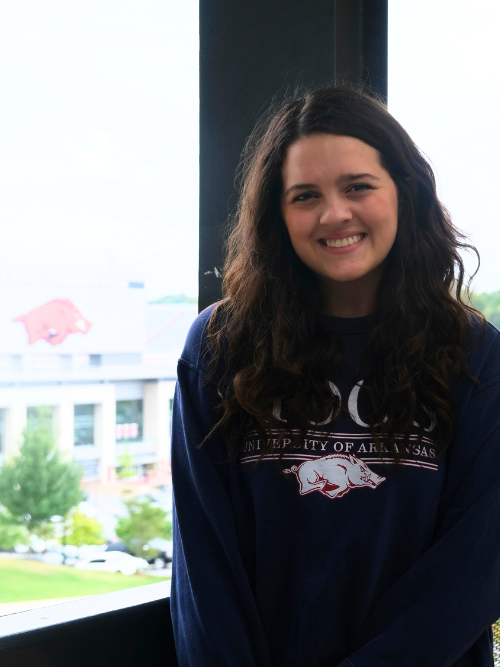 An image of Reese Mitchell, a young white woman with shoulder length brown wavy hair, smiling wide in front of Razorback Stadium