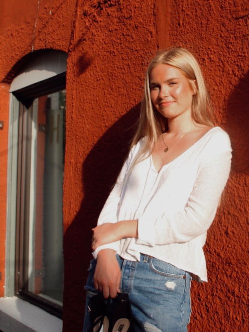 Image of Sierra standing in front of a deep red wall with her hands clasped in front of her, wearing a white blouse.